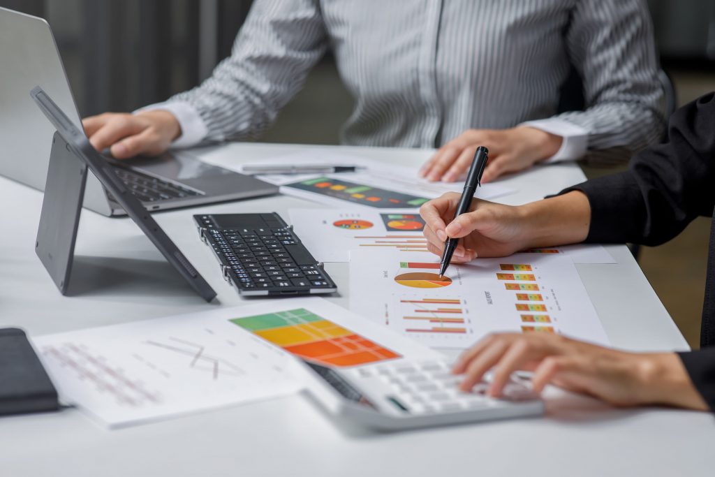 Mãos de duas pessoas brancas fazem anotações sobre papéis de gráficos e fazem cálculos em uma calculadora. 