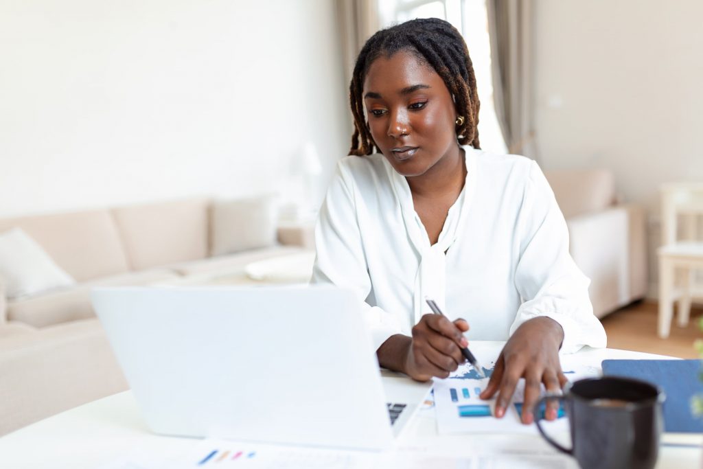 Mulher negra de dreadlocks olha para notebook e faz anotações em um papel com gráficos à frente. 