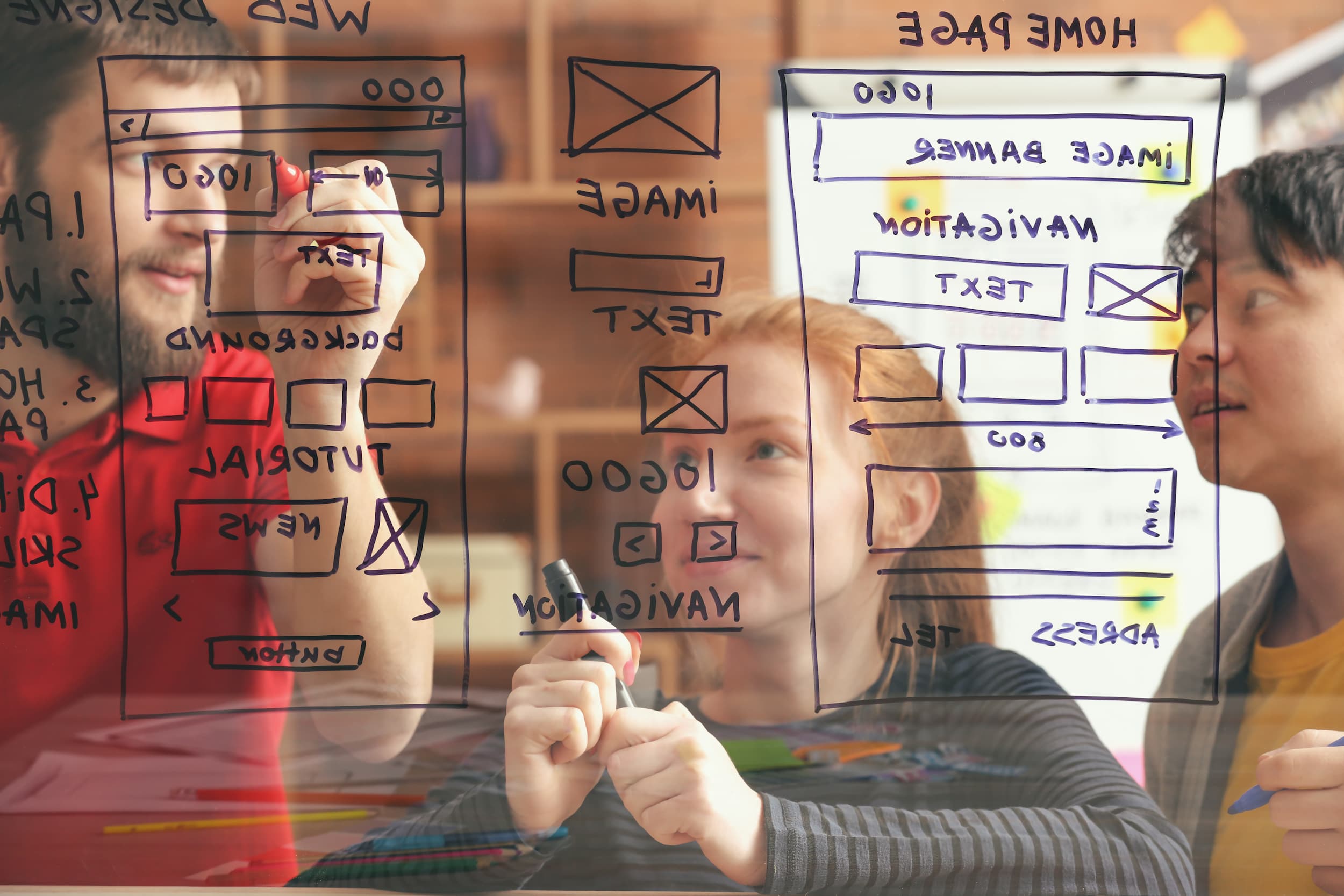 Equipe diversa desenha com canetões desenham um projeto de UI/UX design em quadro transparente.