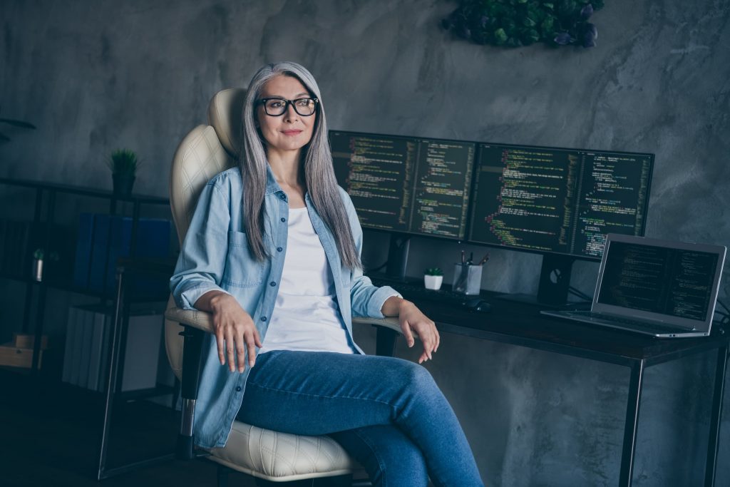 Mulher grisalha de óculos sentada em cadeira, ao lado de diferentes telas de computador com códigos. 