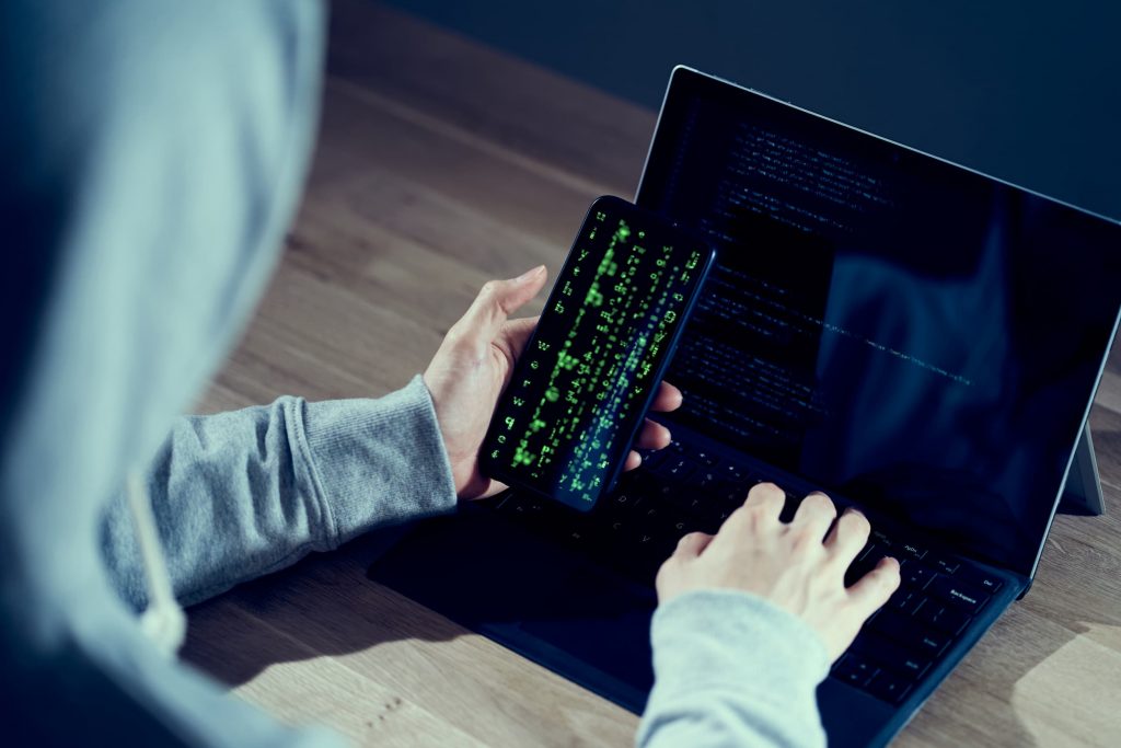 Pessoa hacker de capuz digitando sobre notebook e com um celular na mão, com códigos em verde na tela. 