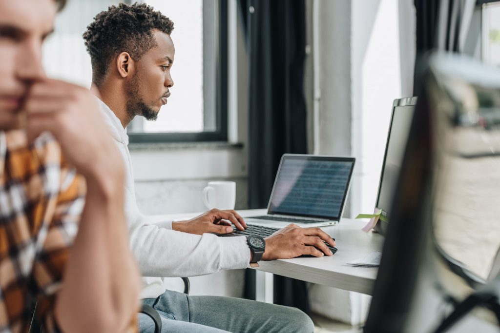 Perfil de um homem negro sentado em mesa de escritório com computador e notebook. Ao lado, em desfoque, um homem branco encara a tela de outro computador.