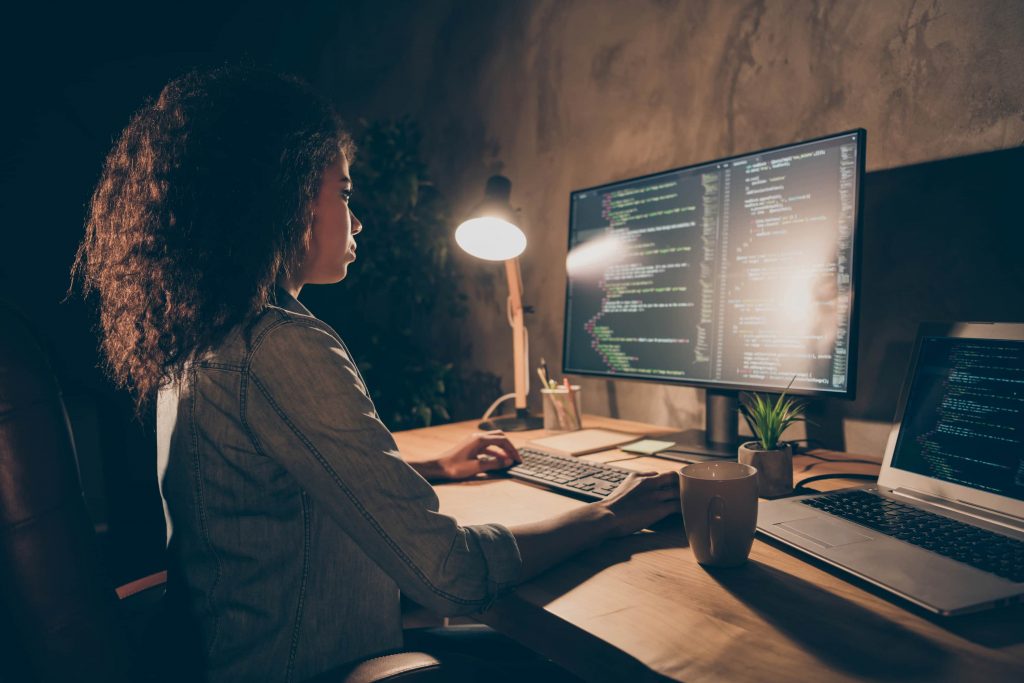 Mulher de cabelos crespos sentada em mesa de escritório programando com dois computadores. 