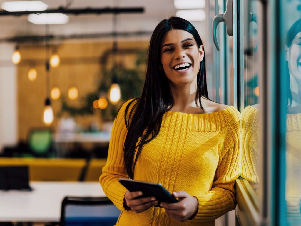  Mulher negra de cabelos lisos em escritório sorrindo enquanto segura um tablet. 