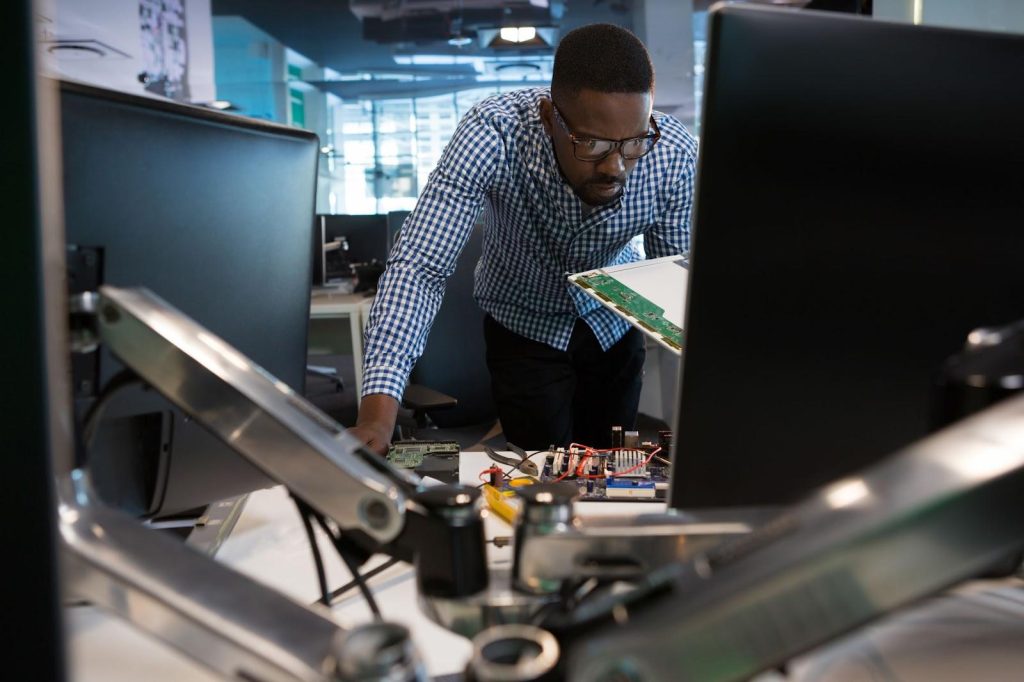 um homem negro de cabelo curto está vestindo roupas sociais claras e usando um óculos de grau enquanto permanece debruçado sobre uma mesa com diversos aparelhos eletrônicos e olha para um destes componentes enquanto segura-o em sua mão