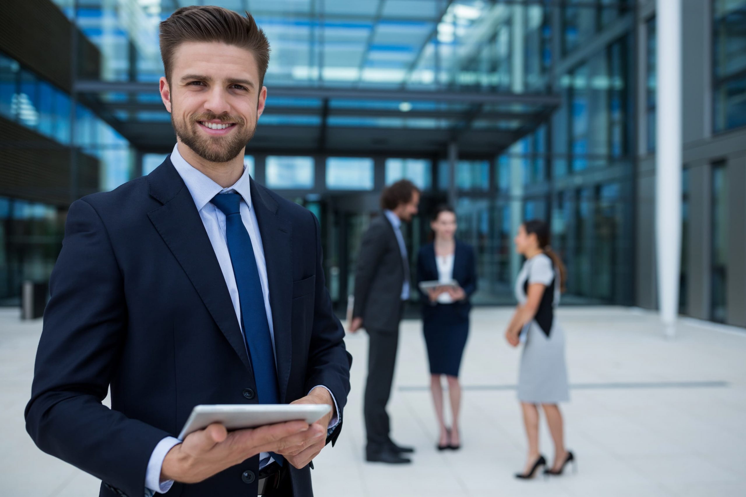Homem branco de terno olha para frente enquanto segura um tablet nas mãos. Ao fundo, em desfoque, diferentes colegas conversam.