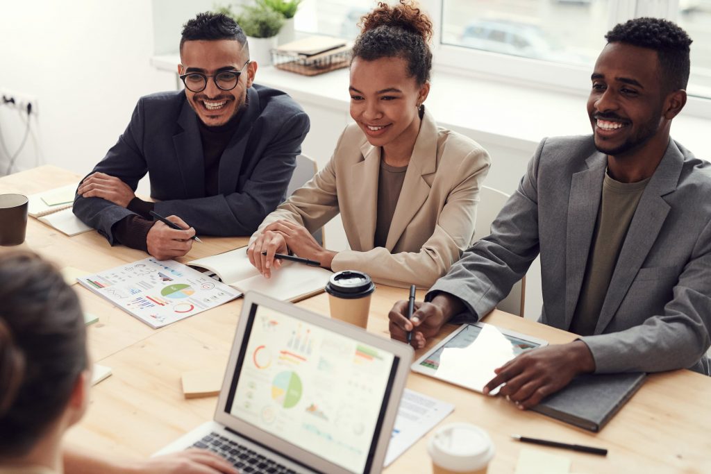 Pessoas diversas sentadas em uma mesa de reunião em escritório, com notebook, tablet e cadernos de anotações à frente. 