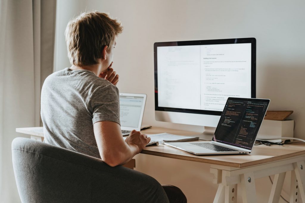 Homem branco de costas sentado em mesa de escritório, com dois notebooks e um monitor à frente.