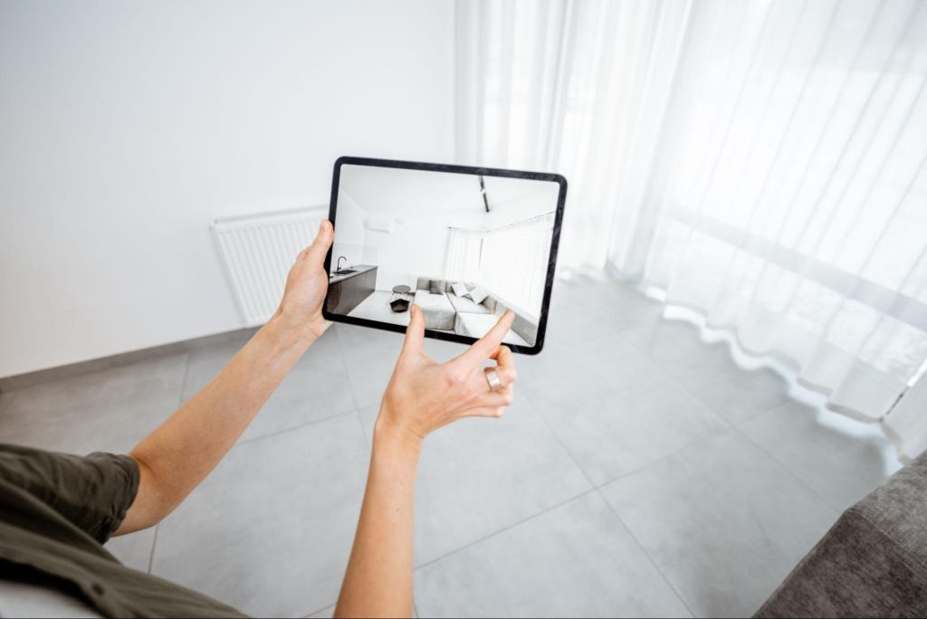 Pessoa com um tablet em mão, com uma imagem de uma sala mobiliada. Ao fundo, um cômodo vazio, dando a entender que a pessoa usa de realidade aumentada.