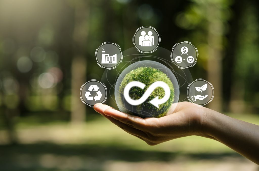 Mão segurando uma bola verde com o símbolo do infinito com uma seta, indicando o reuso. Dela sai uma projeção holográfica de ícones relacionados a TI verde. 