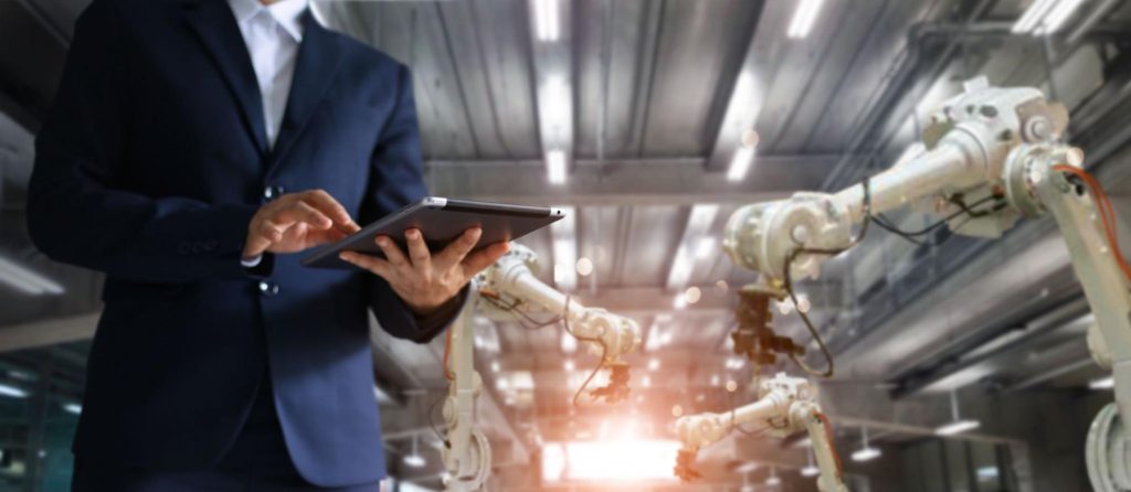 Pessoa de terno, dentro de uma indústria, clica em tablet e movimenta três robôs atrás dela.