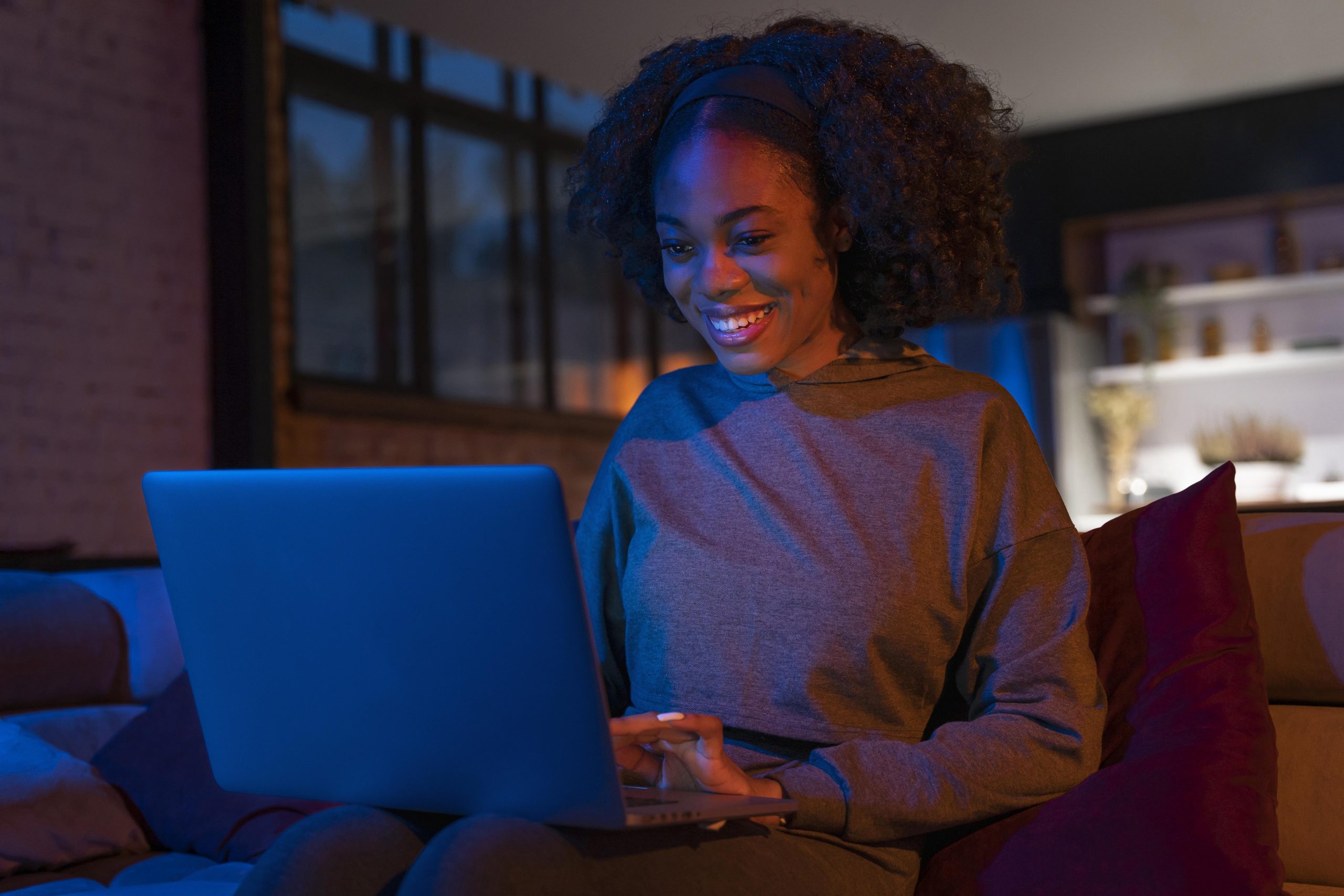 Glossário de cibersegurança: conheça os principais termos! Mulher utilizando notebook, sentada em sofá, com luz azul iluminando o ambiente.