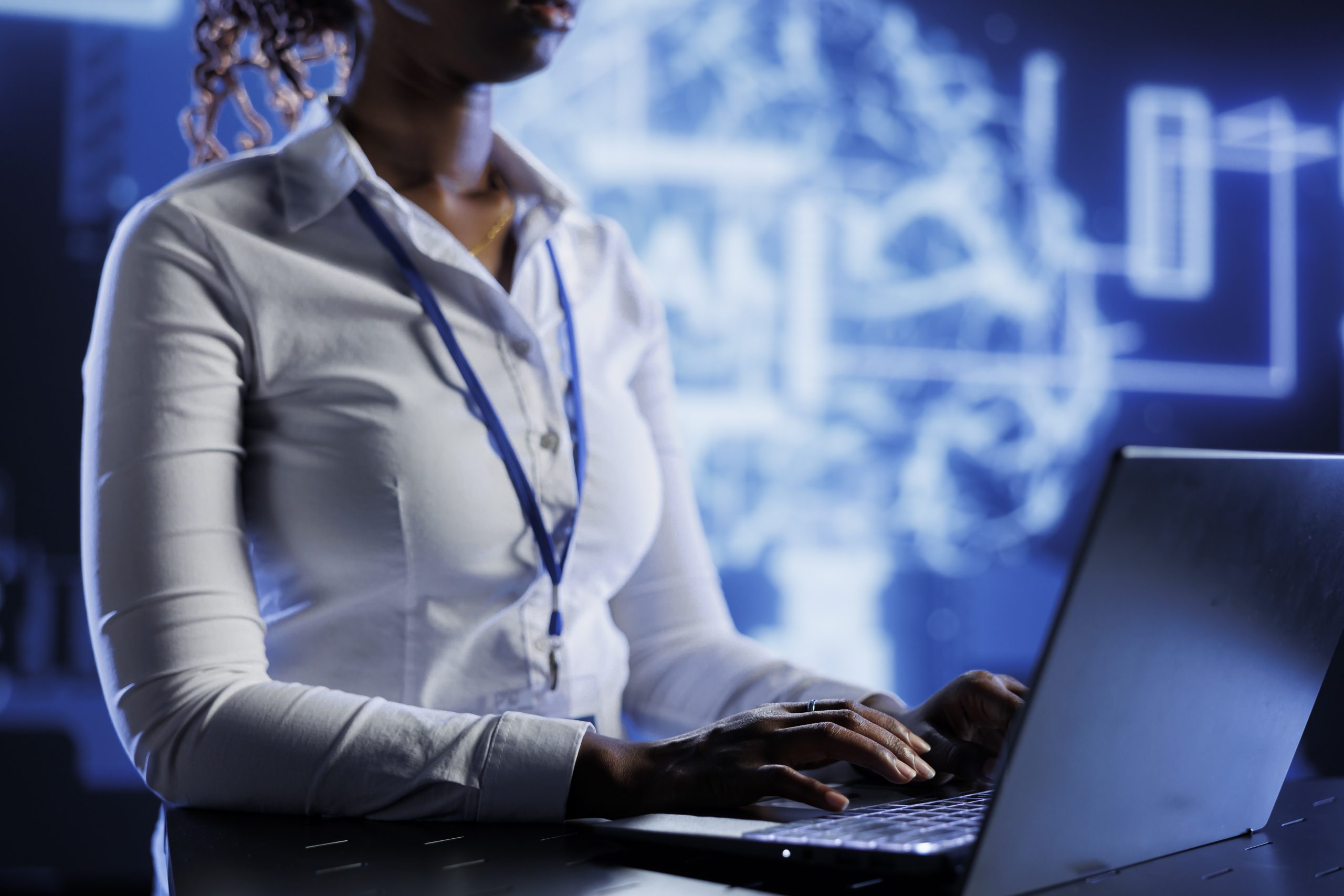 Profissional de TI, vestindo crachá de identificação, utilizando notebook e observando informações presentes na tela.