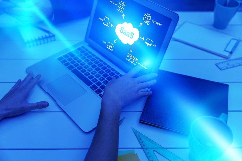 Imagem de tonalidade azul, mostrando pessoa utilizando notebook, enquanto a tela apresenta o texto “SaaS” dentro de uma nuvem, com ícones do universo de TI ao redor.