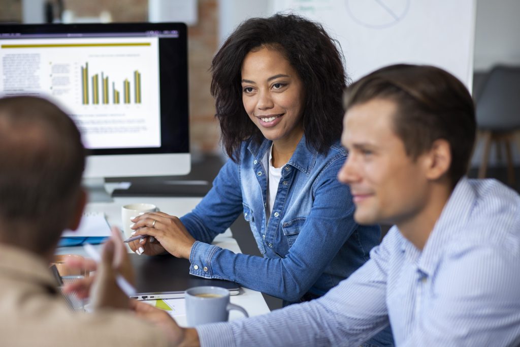 Profissionais da tecnologia em escritório, reunidos em uma mesa com monitores mostrando gráficos e outros dados, com papéis, canecas e demais equipamentos em cima da mesa.