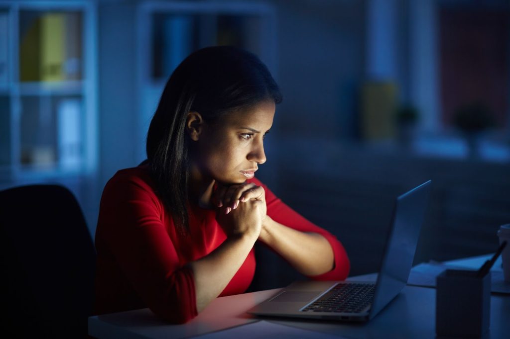 Mulher em frente a notebook, usando blusa vermelha e olhar de preocupação, com janela e armários ao fundo.