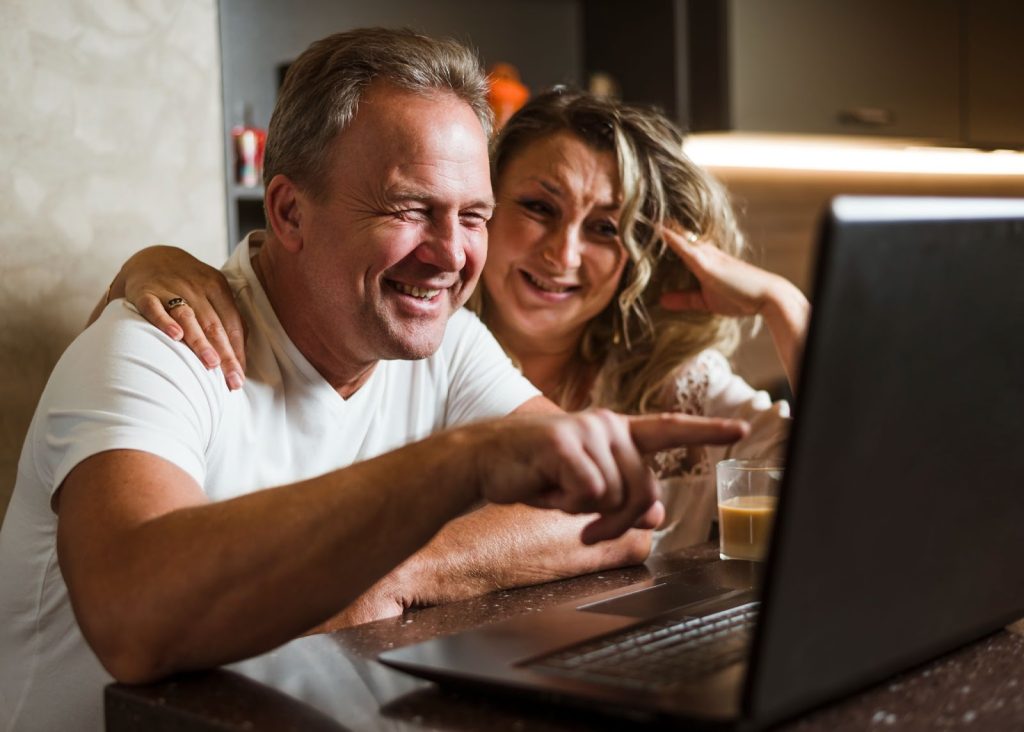 Homem apontando para a tela de notebook, com mulher ao lado e copo com café. Ambos estão sorrindo. O ambiente está bem iluminado.