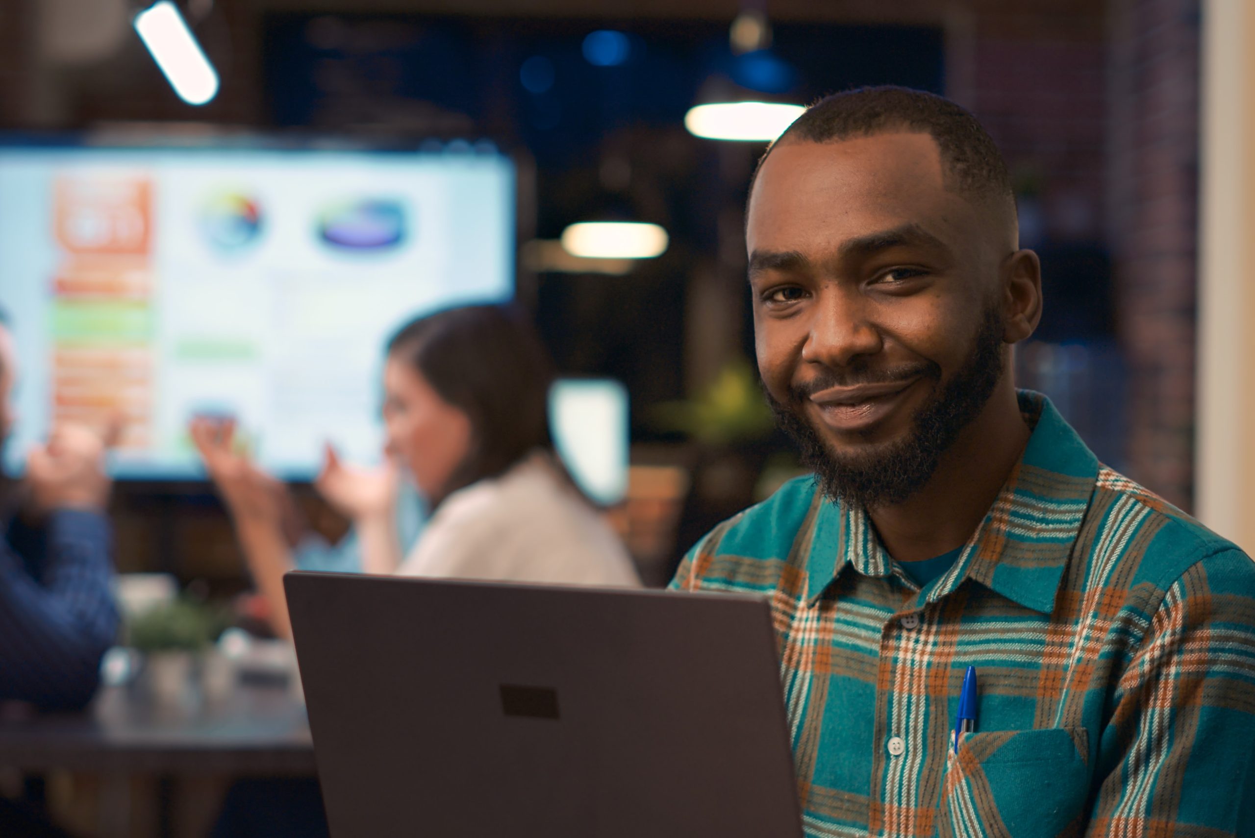 Profissional da tecnologia utilizando notebook, com outros profissionais ao fundo conversando, bem como quadro apresentando dados e gráficos.