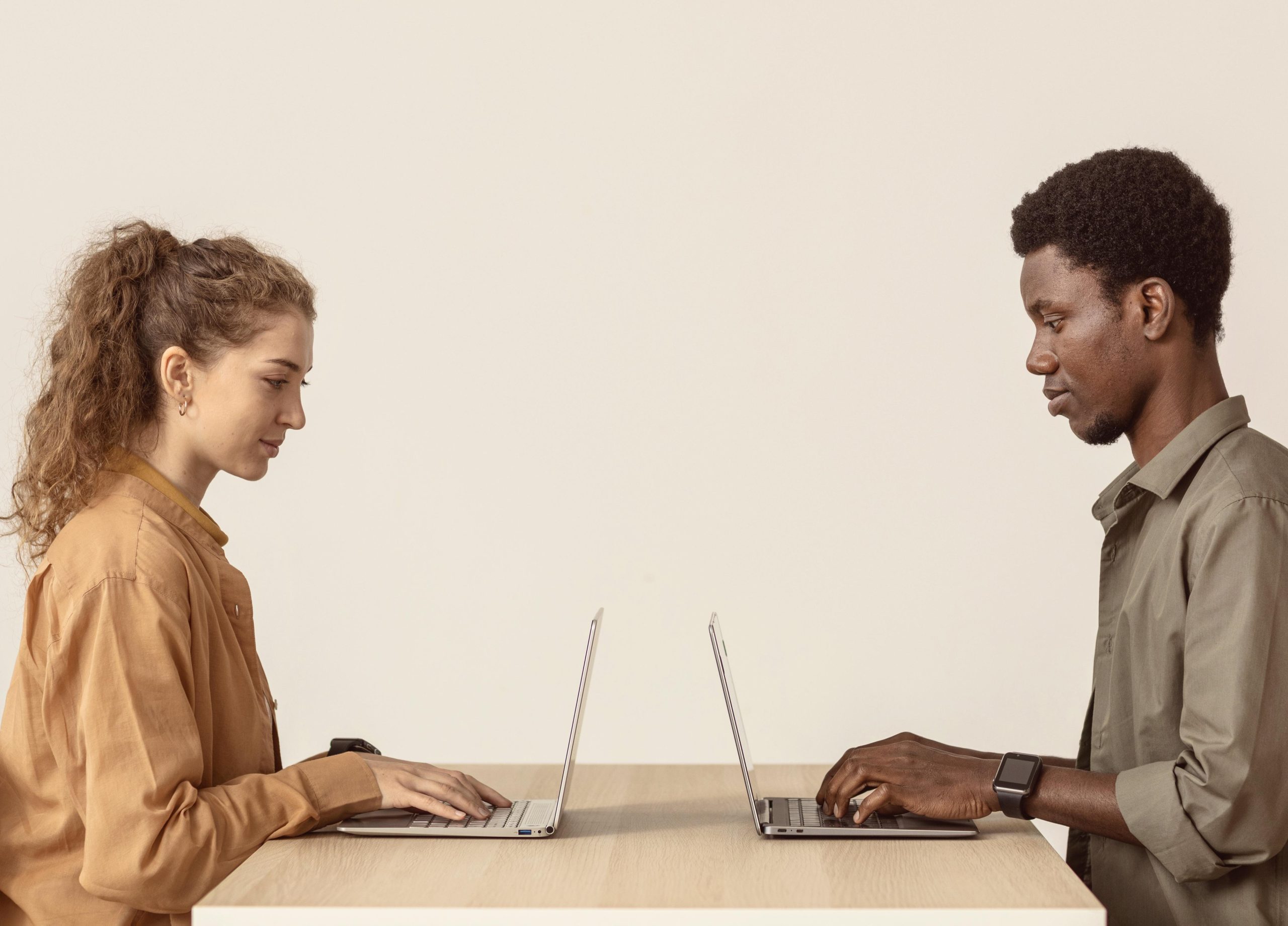 Homem e mulher, sentados um de frente para o outro, ambos utilizando notebooks. Os dois utilizam camisas de botão de tons terrosos. Parede ao fundo branca.