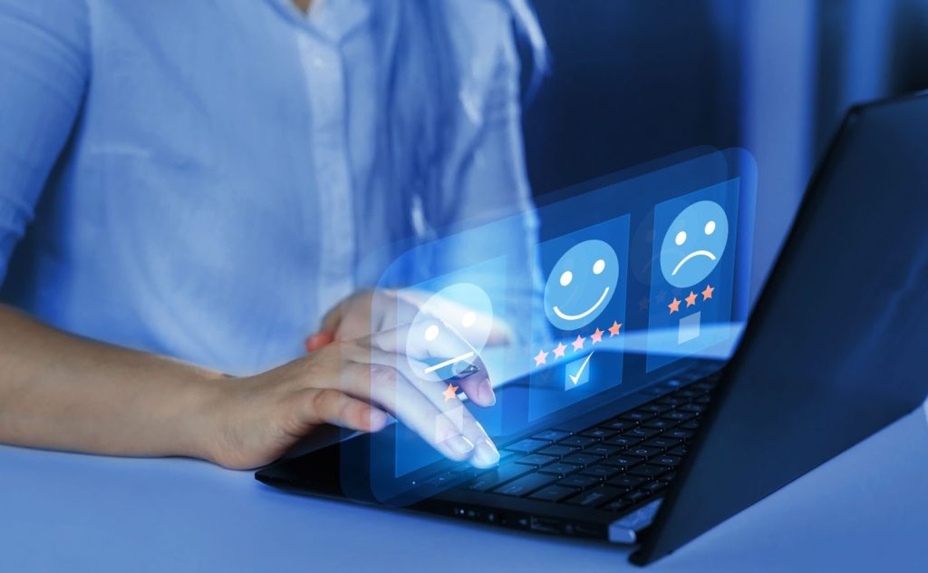 Pessoa utilizando notebook, com imagens que simbolizam uma pesquisa de satisfação. Estes símbolos parecem sair da tela.