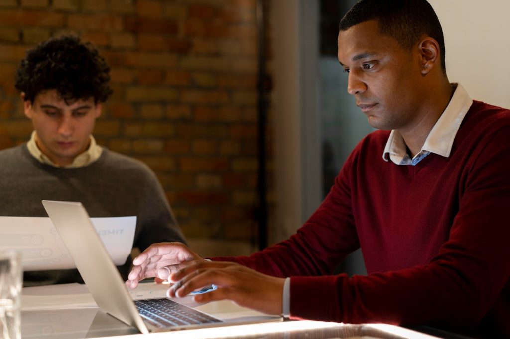 Profissionais da tecnologia utilizando notebook e lendo papel com informações. Ao fundo, parede de tijolos e um copo está acima da mesa.