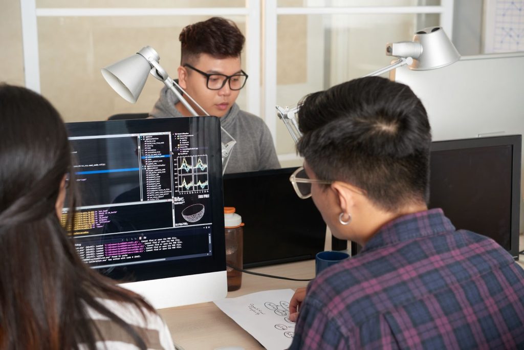 Profissionais de TI utilizando computadores que apresentam dados na tela. Em cima da mesa estão canecas, garrafa, luminárias e papéis.