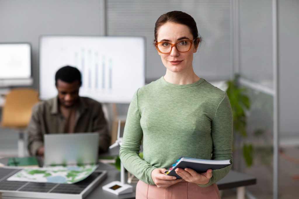 Profissionais da tecnologia, mulher a frente segurando caderno e homem ao fundo utilizando notebook. Na mesa, tela de energia solar e imagens representando reservas ambientais. Ao fundo, telas demonstrando dados gráficos e plantas. Representação da sustentabilidade empresarial.
