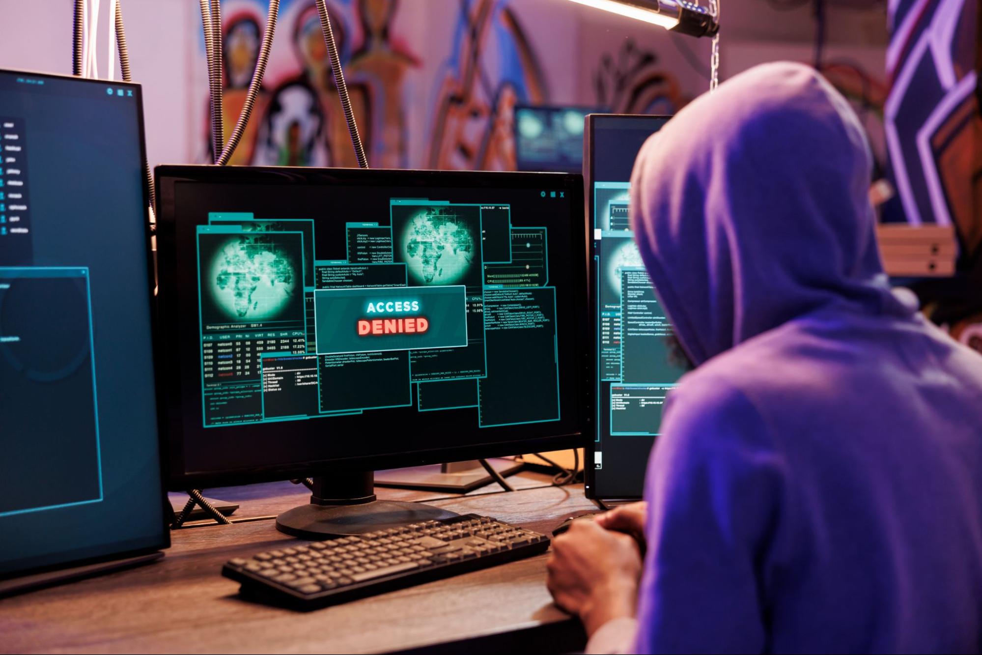 Pessoa utilizando computador, possivelmente um hacker, com imagens na tela e o escrito “Acesso negado”, em inglês. O indivíduo utiliza capuz roxo e, ao fundo, há uma parede com desenhos.