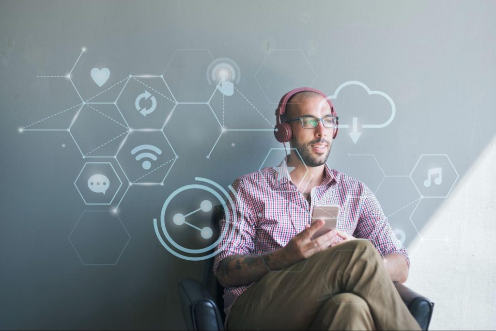 Homem sentado em poltrona, com pernas cruzadas, utilizando fone de ouvido e segurando celular. Por cima da imagem, símbolos de tecnologia, como wi-fi e nuvem, aparecem interligados por linhas.