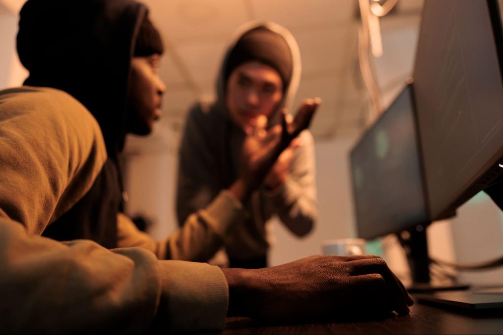 Dois profissionais da tecnologia utilizando computador, com dois monitores, enquanto conversam. A imagem está levemente embaçada e, ao fundo, há uma parede branca.