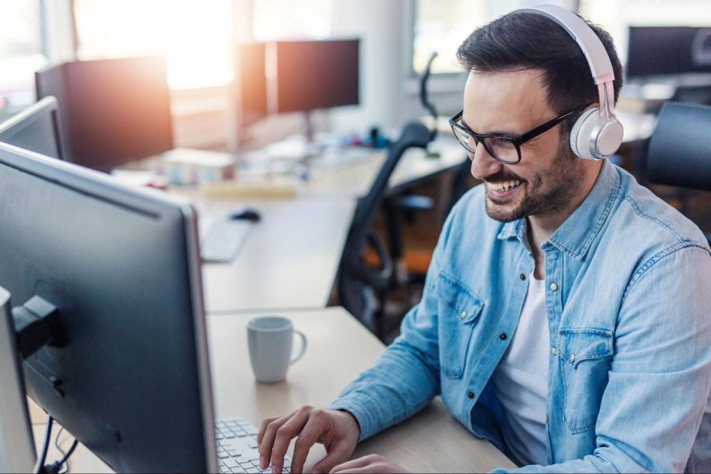  homem branco vestindo camisa jeans sorri enquanto olha para o monitor de um computador, que está em cima de uma mesa de madeira clara