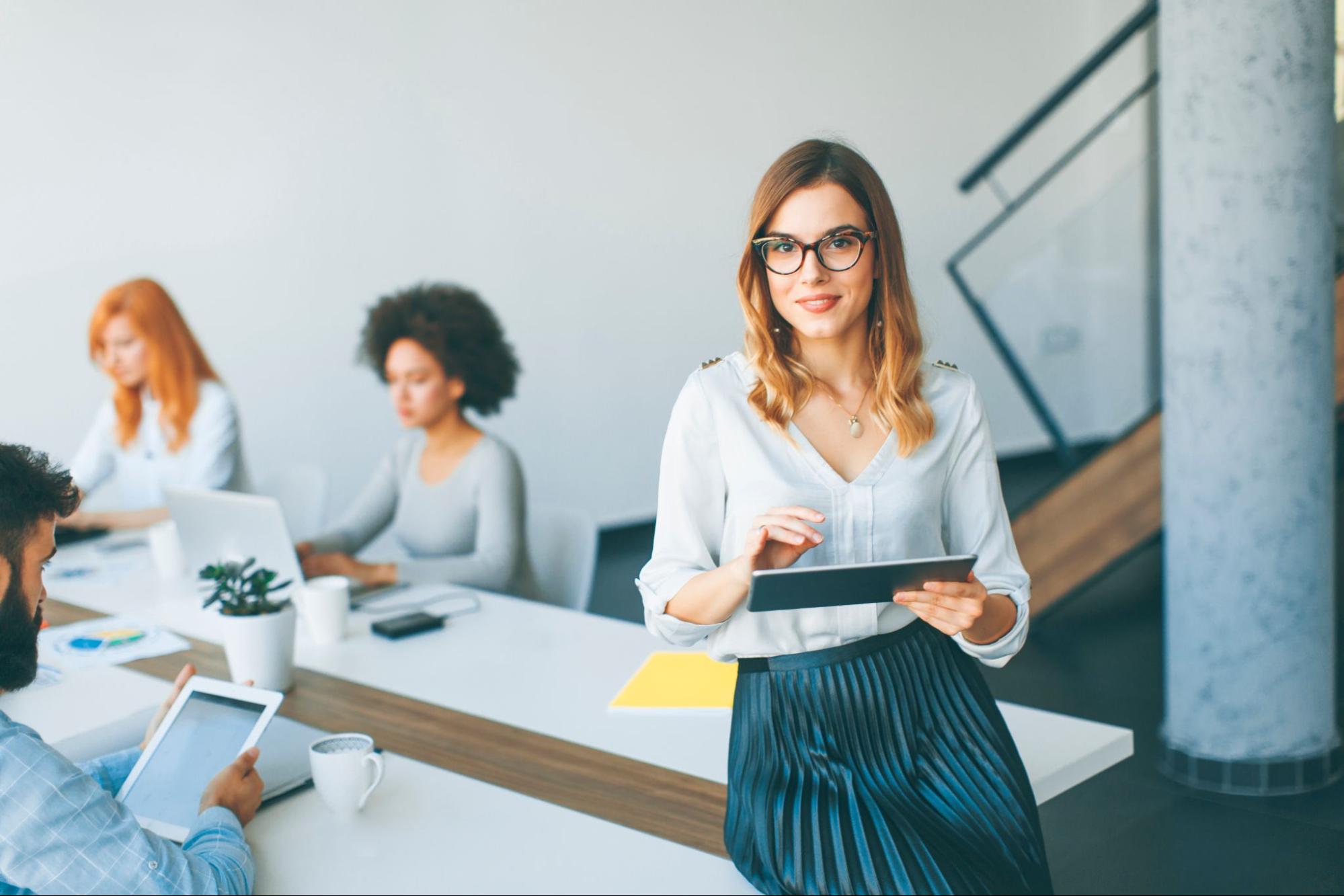 linhas de crédito para empresas mulher vestindo saia preta plissada e camisa branca social está parcialmente sentada em bancada branca, num ambiente de escritório em que três mulheres permanecem sentadas, digitando em seus notebooks.