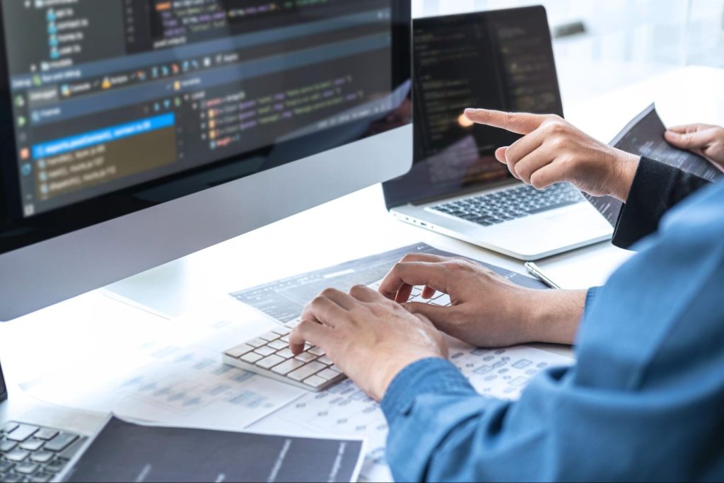 profissionais de qualidade de software apontando para telas de computador e digitando em notebook enquanto permanecem sentados em frente a mesa clara que comporta os aparelhos