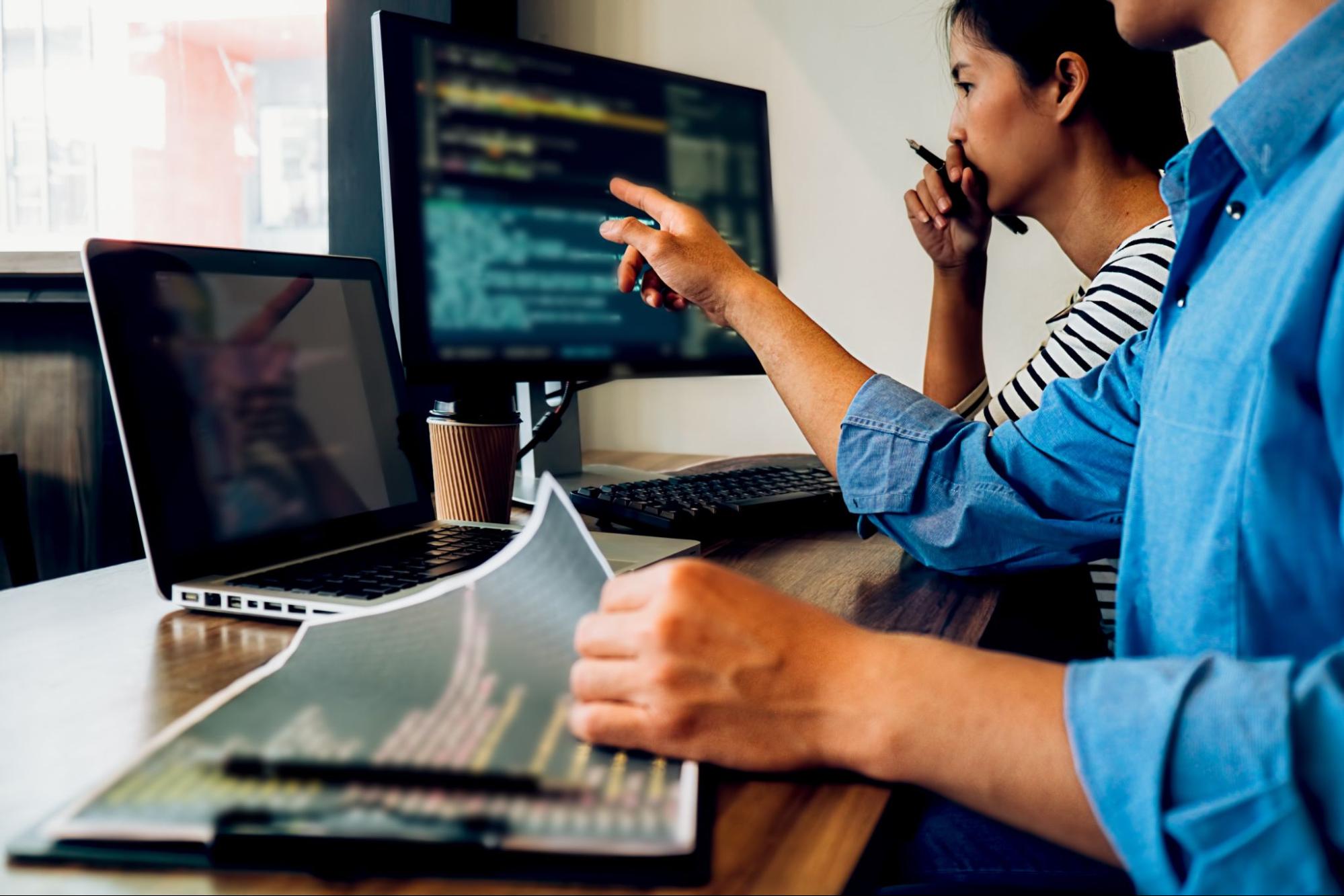 image3 profissionais de qualidade de software apontando para telas de computador e notebook enquanto permanecem sentados em frente a mesa de madeira que comporta os aparelhos