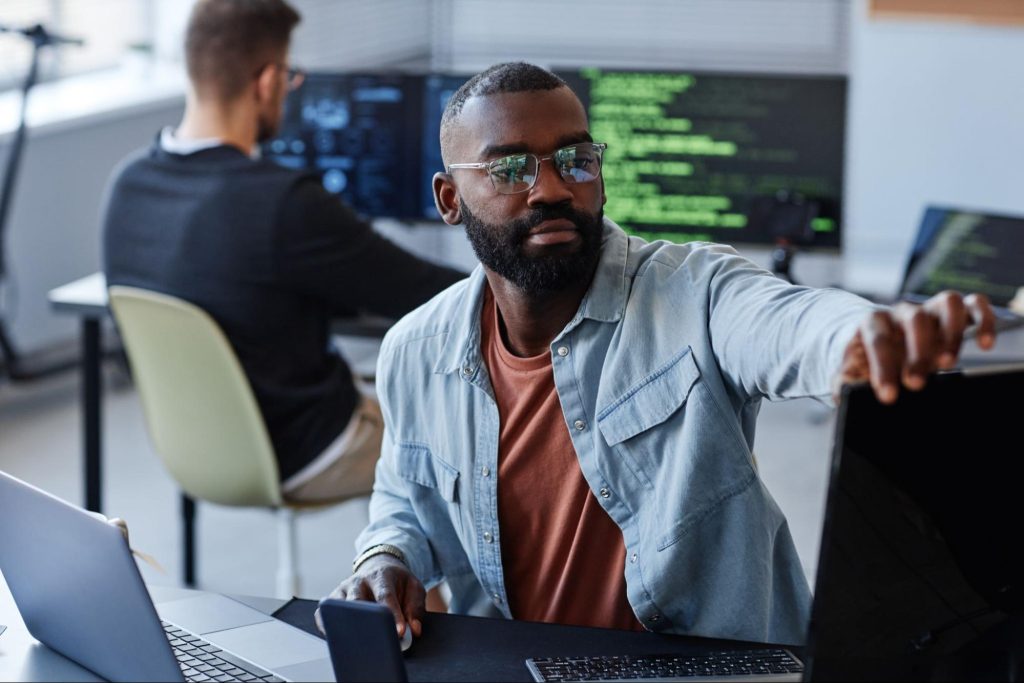 Um homem negro, usando óculos e uma camisa azul clara, está sentado em uma mesa de trabalho. Ele parece concentrado enquanto estende a mão para ajustar um monitor. Ao fundo, outro homem está sentado de costas, trabalhando em um computador com telas que exibem códigos. O ambiente é moderno e bem iluminado.