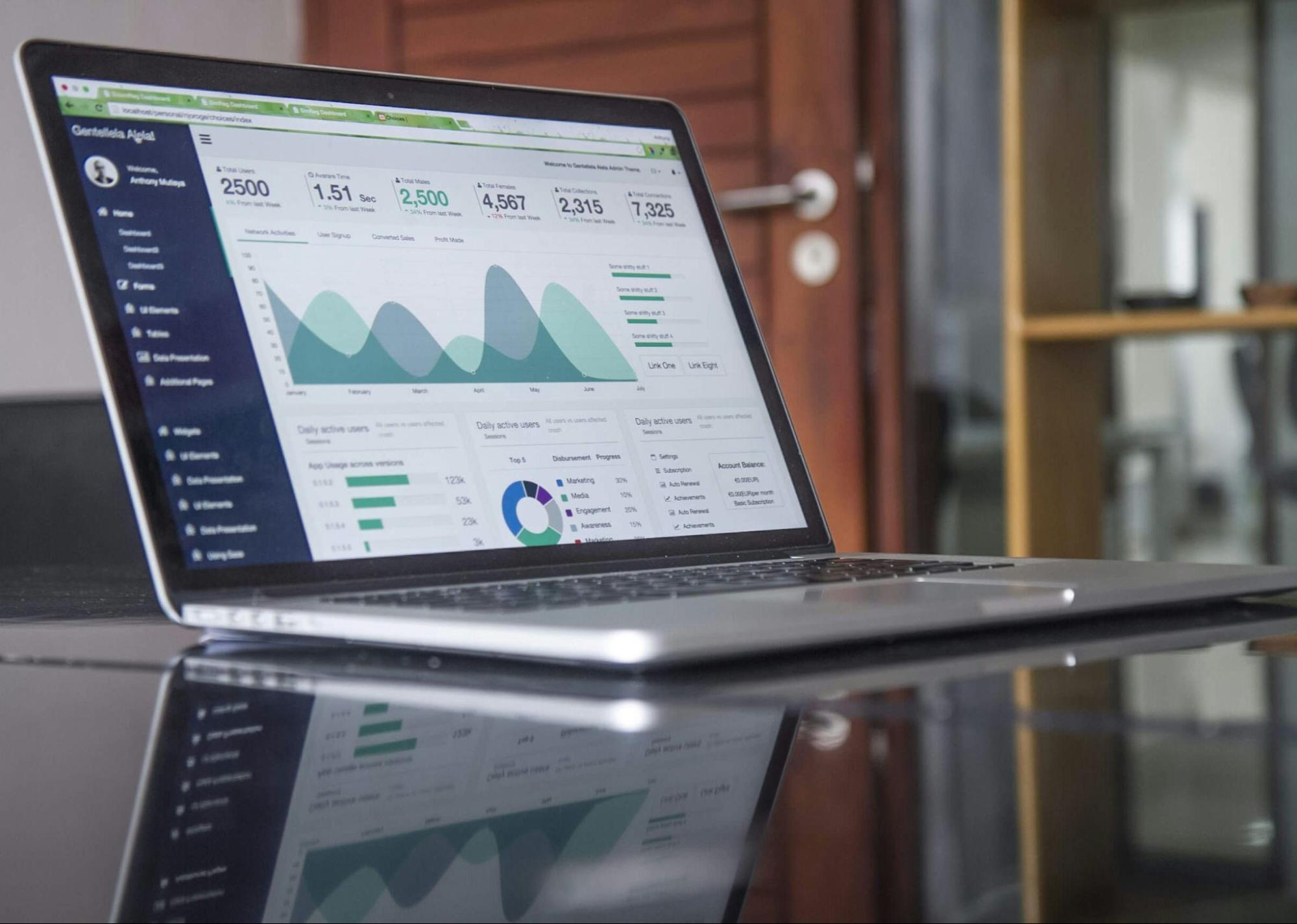 Laptop exibindo um painel de controle com gráficos e dados analíticos, refletido em uma superfície escura. O fundo apresenta uma porta de madeira e prateleiras.