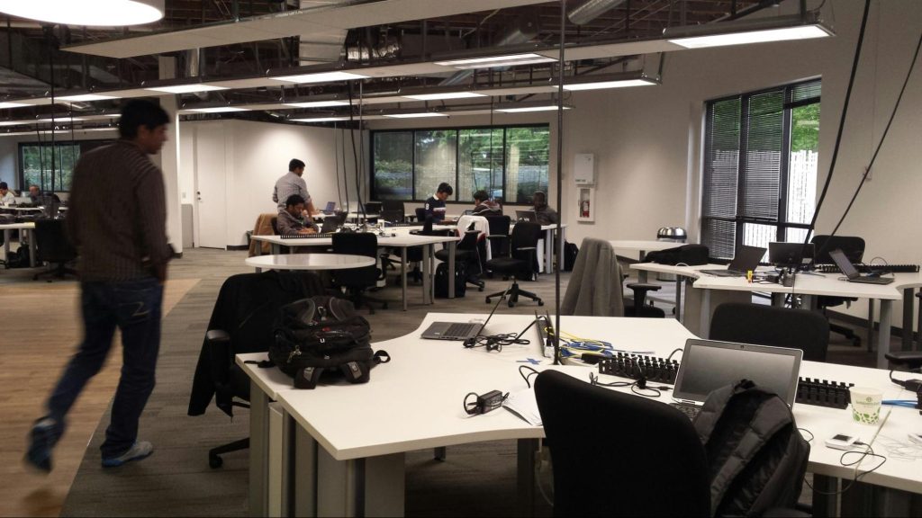 Espaço de trabalho com várias mesas e profissionais em atividade. Alguns estão sentados em frente a laptops, enquanto outros estão em pé ou em movimento. A iluminação do ambiente é clara, e o design é contemporâneo.