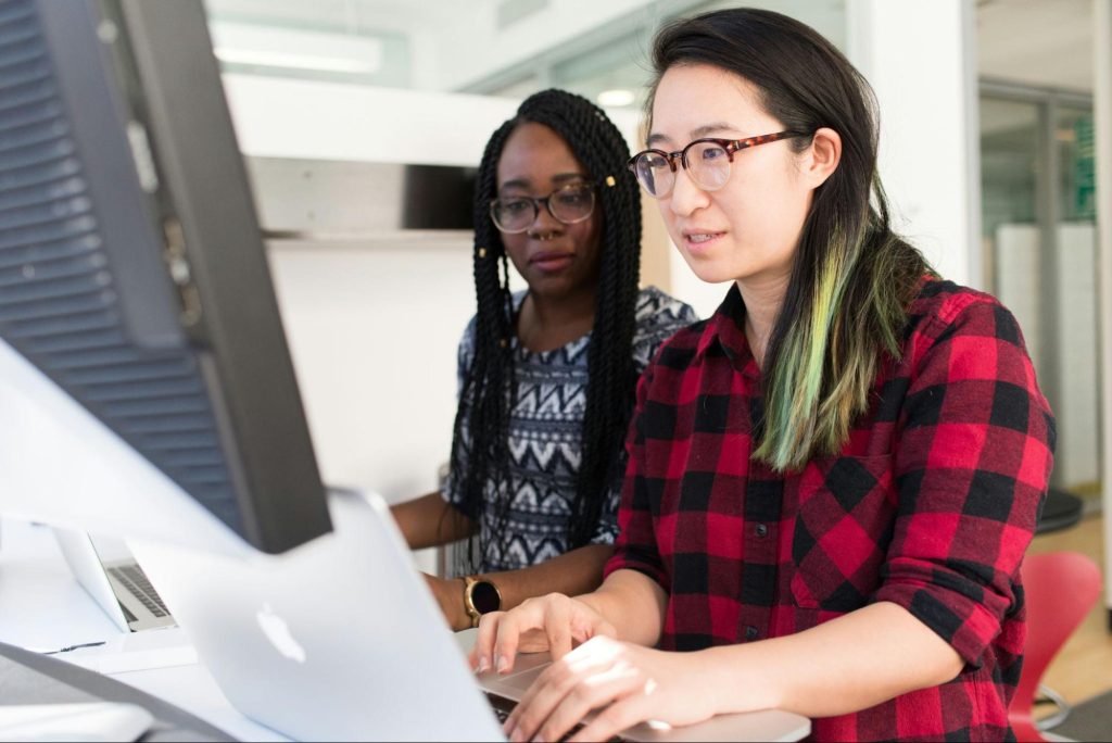 Duas mulheres estão trabalhando juntas em um computador. A mulher à direita, com cabelo liso e mechas verdes, está digitando em um laptop. A mulher à esquerda, com tranças e óculos, observa atentamente a tela. Ambas parecem concentradas na tarefa. O ambiente é claro e moderno, com mesas brancas ao fundo.