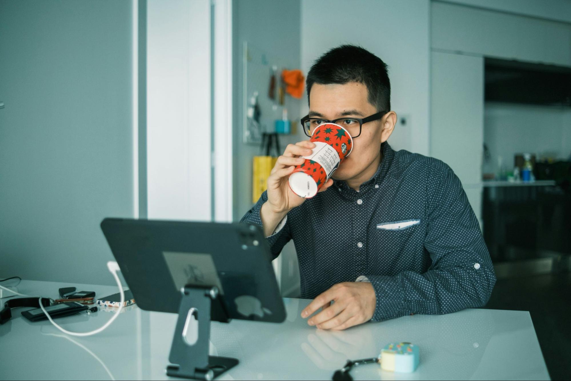 : Homem vestindo  camisa cinza e óculos de grau está tomando café enquanto olha para tablet em cima de mesa clara