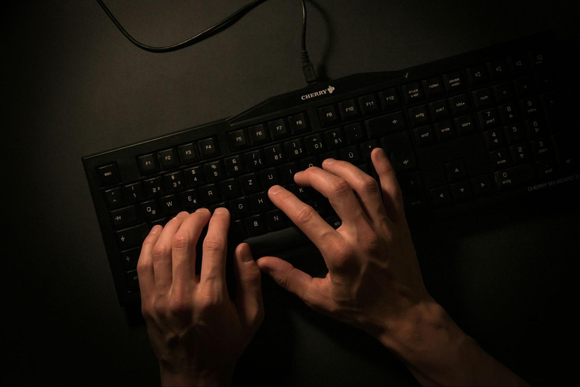 mãos humanas digitam sobre teclado preto com algarismos e letras brancas, disposto sobre superfície escura.