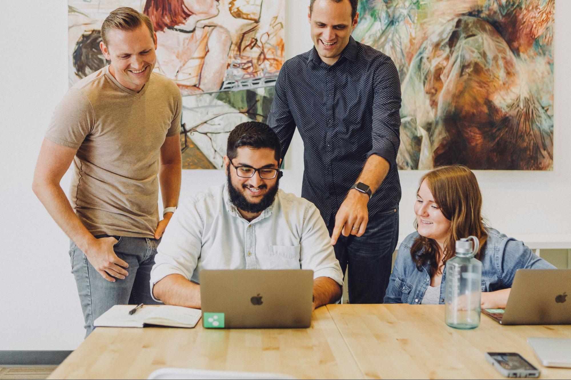 o-que-e-ciberseguranca Grupo de quatro pessoas em uma mesa, interagindo e sorrindo enquanto observam um laptop. Um homem de camisa clara está sorrindo para a tela, enquanto outros três, em pé, discutem algo com entusiasmo. Ao fundo, há uma obra de arte colorida na parede.