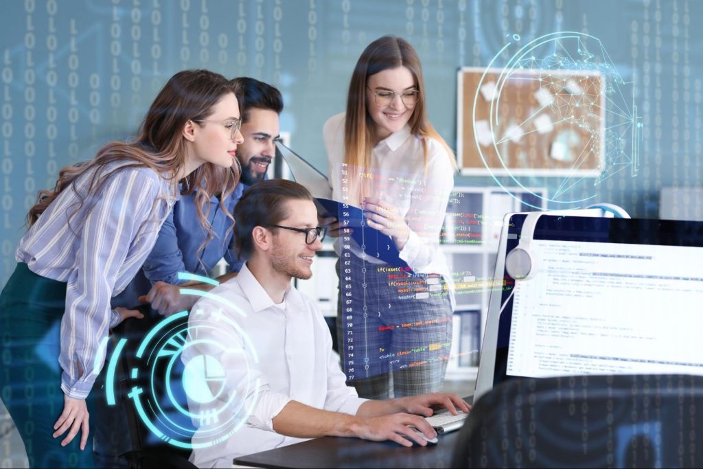 Grupo de pessoas em uma reunião, um homem apresentando para os outros ao lado de um computador. Post-its e material de escritório estão visíveis na mesa.